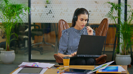 Indian young adult gen z busy woman lady sitting on chair wear headphone use laptop look screen do...