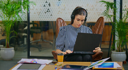 Indian young adult gen z busy woman lady sitting on chair wear headphone use laptop look screen do...