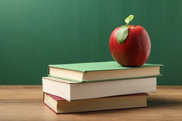 Different books and red apple on wooden table near blackboard, closeup