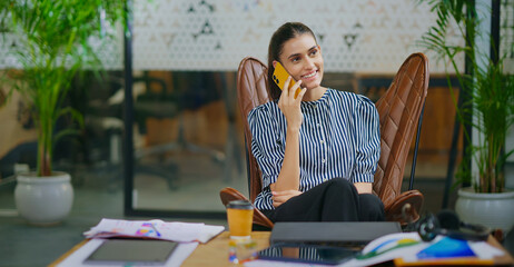 Indian young adult gen z woman lady sitting on chair hold mobile talk gossip enjoy call indoor job...