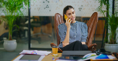 Indian young adult gen z woman lady sitting on chair hold mobile talk gossip enjoy call indoor job work place happy female girl expert staff use phone speak have fun joy spend day time break at office