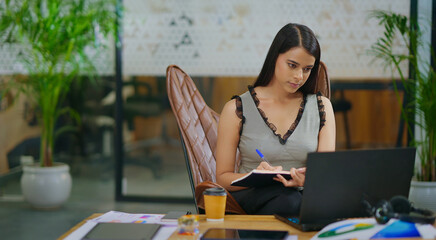 Indian gen z busy woman lady sitting chair use laptop on desk table look screen check email job...