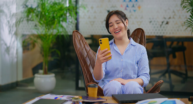 Afghan young adult gen z woman lady sitting on chair use mobile look cam talk gossip indoor job place happy girl staff hold phone speak on video call after work enjoy break spend day time at office - Powered by Adobe
