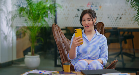 Afghan young adult gen z woman lady sitting on chair use mobile look cam talk gossip indoor job place happy girl staff hold phone speak on video call after work enjoy break spend day time at office