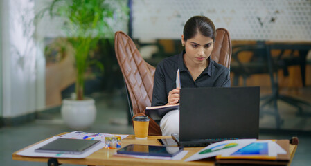 Indian gen z busy woman lady sitting chair use laptop on desk table look screen check email job...