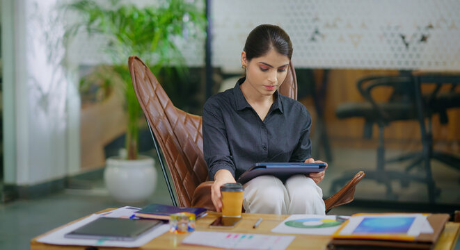 Indian young adult gen z busy woman use tablet device look screen do online study data work teen girl lady female intern staff pick up paper coffee cup during day break time at job office place