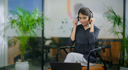 Indian happy young adult gen z busy woman lady girl wear headphone sitting on chair play online mp3...