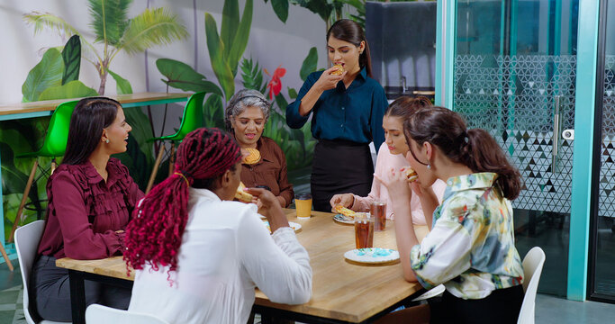 Happy asian woman girl old lady senior group team talk gossip eat sandwich share lunch indoor office work place Indian diverse female sitting around table enjoy breakfast tasty juice cup have fun joy