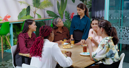 Happy asian woman girl old lady senior group team talk gossip eat sandwich share lunch indoor office work place indian diverse female sitting around table enjoy breakfast tasty juice cup have fun joy
