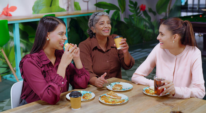 Indian happy senior woman lady group sitting around table eat break time tasty lunch share food hold cup drink coffee work place female girl staff talk gossip enjoy day have fun joy job office
