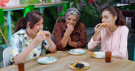 Indian happy senior woman lady group sitting around table eat break time hold tasty piece lunch share food work place female girl staff talk gossip enjoy day have fun joy job office