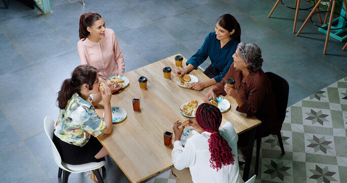 Indian happy woman staff sitting around table with old aged lady talk gossip eat tasty pizza junk food enjoy break time female girl group team speak funny joke drink coffee have fun joy at work place