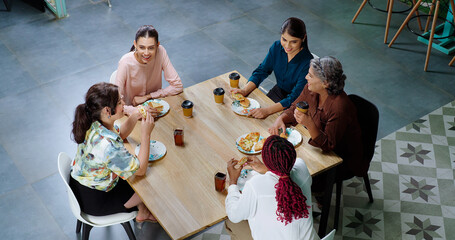 Indian happy woman staff sitting around table with old aged lady talk gossip eat tasty pizza junk food enjoy break time female girl group team speak funny joke drink coffee have fun joy at work place