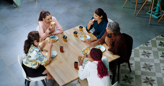 Indian happy woman staff sitting around table with old aged lady talk gossip eat tasty pizza junk food enjoy break time female girl group team speak funny joke drink coffee have fun joy at work place