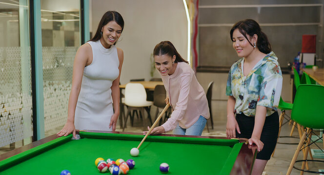 Indian happy young adult gen z woman lady hold cue stick hit ball play sport game on pool table after work girl female staff enjoy good break spend day time have fun joy job place