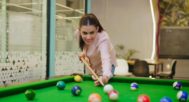 Indian young adult gen z happy woman lady hold cue stick play snooker game on table pool after work at indoor office. Asian girl staff intern enjoy casual break spend good time have fun joy job place