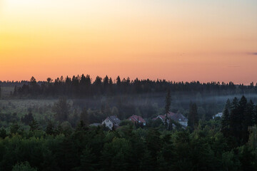 Misty dawn over small village houses surrounded by forest at golden sunrise