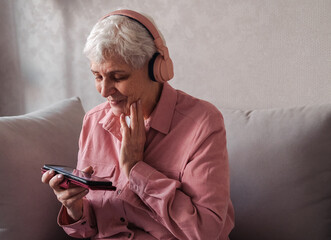 Smiling relaxed senior woman wearing headphones sitting on sofa at home using smart phone looking...