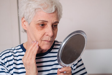 Woman Looking At Herself In The Mirror. Beautiful elderly woman holding mirror and applying face cream at home.