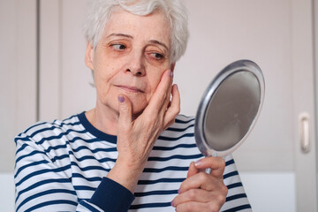 Senior elderly female with gray hair and wrinkles looking in mirror, upset with aging process of her body, reflecting upon her youth and young facial skin, missing her past beautiful appearance