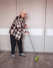 An elderly woman wearing casual clothes is sweeping the room.