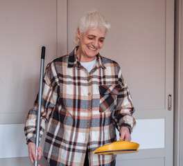An elderly woman wearing casual clothes is sweeping the room.