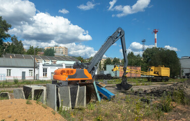 Orange excavator on construction site with concrete blocks and yellow crane