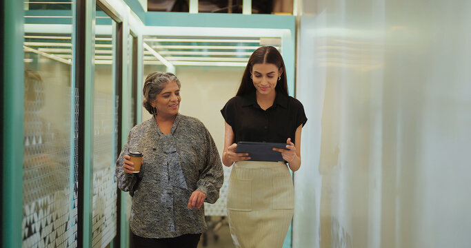 Indian older woman lady talk gossip young adult staff discuss work day time task indoor office place smiling female hold paper cup drink coffee expert girl hold tablet device walk at hall way space