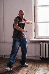 A 40-year-old bald man in casual clothes dances in a room next to a window.