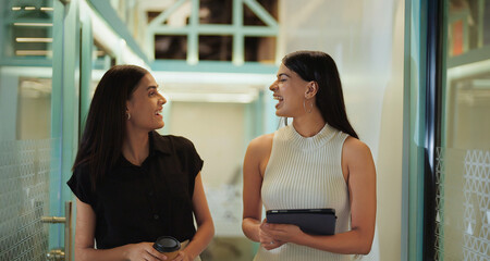 Indian young adult gen z woman lady use tablet walk office space talk work task two expert girl staff hold paper cup drink coffee speak casual enjoy break spend day time at job place have fun joy