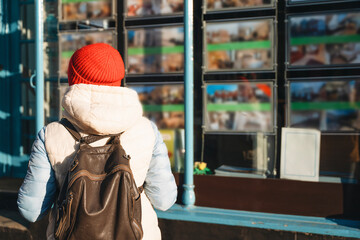 Young person with a red hat observes display boards filled with images of properties in a bustling city during bright daylight