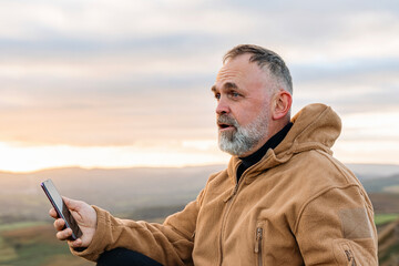 Man in a brown jacket using smartphone while sitting outdoors against a scenic sunset backdrop in the countryside