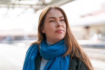 Woman with long hair wearing a blue scarf looks thoughtfully at the train station during a sunny...