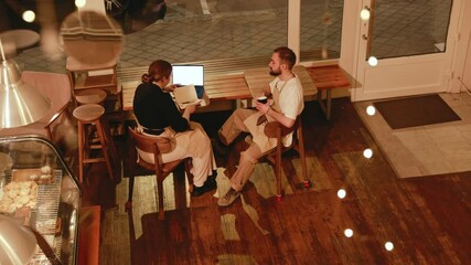 Male employee holding a cup and talking to female owner who is typing on laptop and writing in notebook while they sit at tables