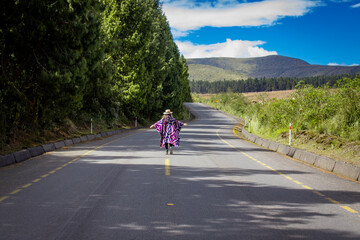Woman wearing a vibrant poncho and hat walks along a deserted road lined with lush green trees,...