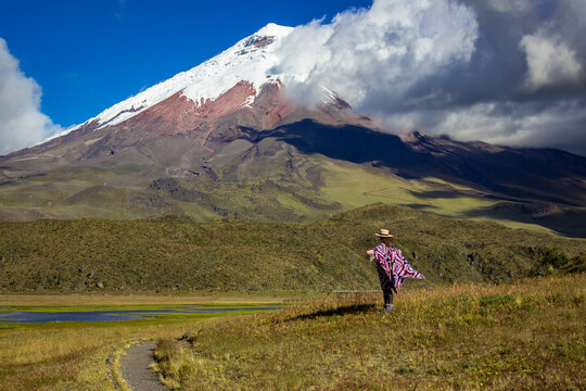 Woman in traditional attire stands admiring the breathtaking view of cotopaxi volcano. The scene captures the vibrant contrast between lush greenery and the snow-capped peak
