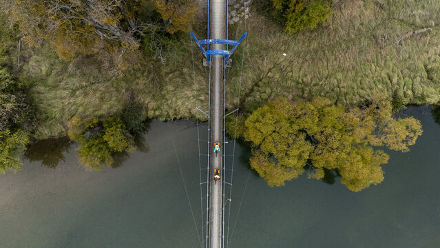 Aerial view of cyclists gliding across the slender CB Kidson bridge, flanked by the river's dark waters and vibrant trees, Waimea River, Tasman Region, New Zealand.