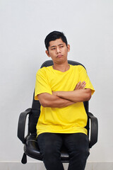 An Asian man sits in an office chair looking bored and with arms crossed over his chest