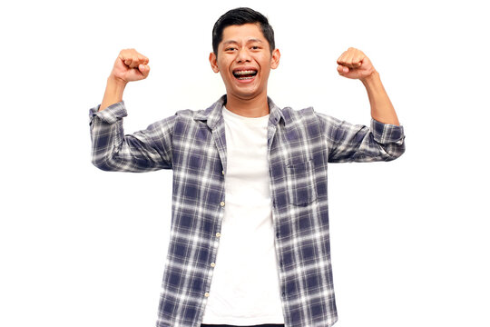 A young Asian man celebrates success with a joyful expression and clenched fists on a white background