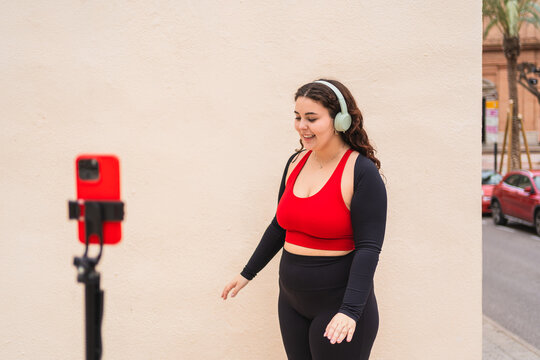 Curvy woman wearing sportswear and headphones recording online fitness class with smartphone on tripod - Powered by Adobe