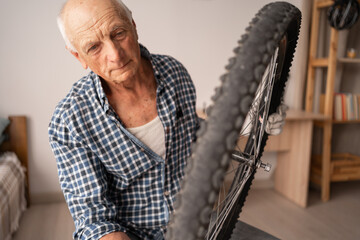 Elderly mechanic repairing bicycle holding cycle wheel in hands sitting on chair in living room....