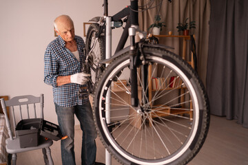 Senior man repairing bicycle in living room preparing for summer. Concept of indoor transport care,...