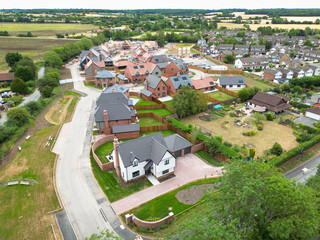 Aerial view of a large, newly completed white detached family home seen at the entrance to a British housing development site in rural Essex. The original village is on the right of the new estate.