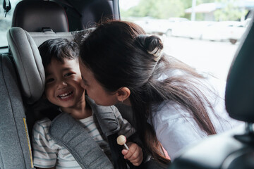 Mother kissing smiling toddler in car seat while he holds a lollipop. Parental care and safety, Family readiness before traveling.