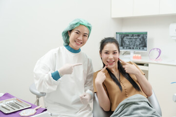 Asian female dentist adult pointing toward female patient adult sitting on dental chair smiling...