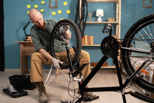 Young bearded man fixing wheel while repairing bicycle at home