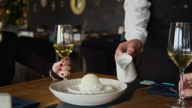 Cropped shot of unrecognizable waiter in black and white uniform pouring liquid nitrogen onto dome dessert creating smoky effect at fine dining table