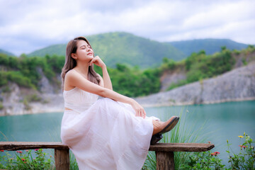 asian woman wearing white dress sitting relax on wooden table by the lake and mountain nature...