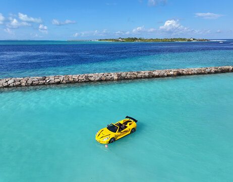 Aerial view of a striking yellow car floating on the aquamarine waters near the rocky barrier, with the distant island a contrast of green against the deep blue sea, Thulusdhoo, Kaafu Atoll, Maldives.