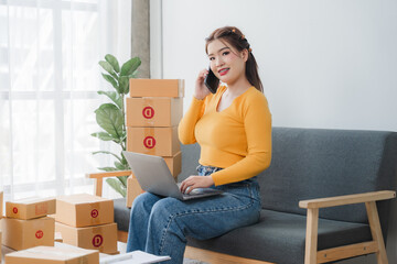 E-commerce Hustle: Young Asian entrepreneur managing orders and logistics on phone and tablet, surrounded by shipping boxes in her home office.	